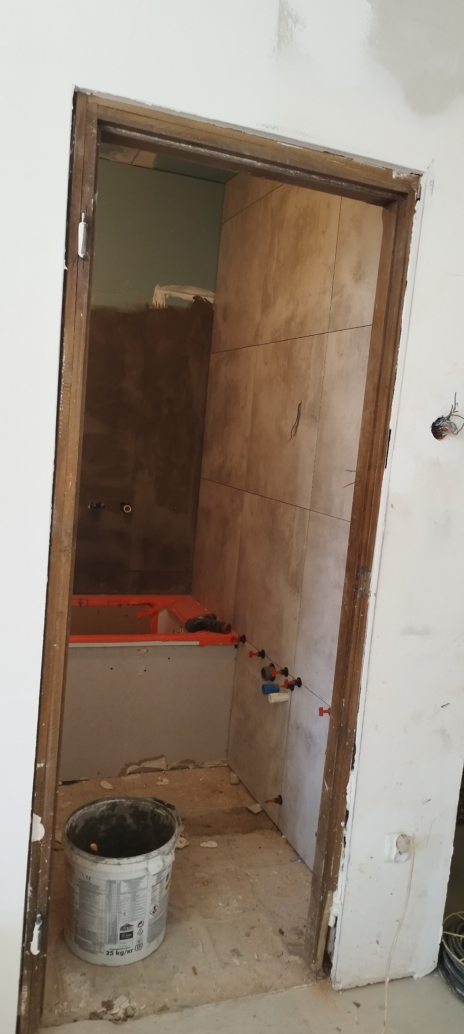 Łazienka w trakcie remontu: widok przez futrynę na częściowo wyłożoną beżowymi płytkami ścianę, z widocznymi elementami instalacji wodnej i poziomowania płytek, oraz wiadro po zaprawie na podłodze.