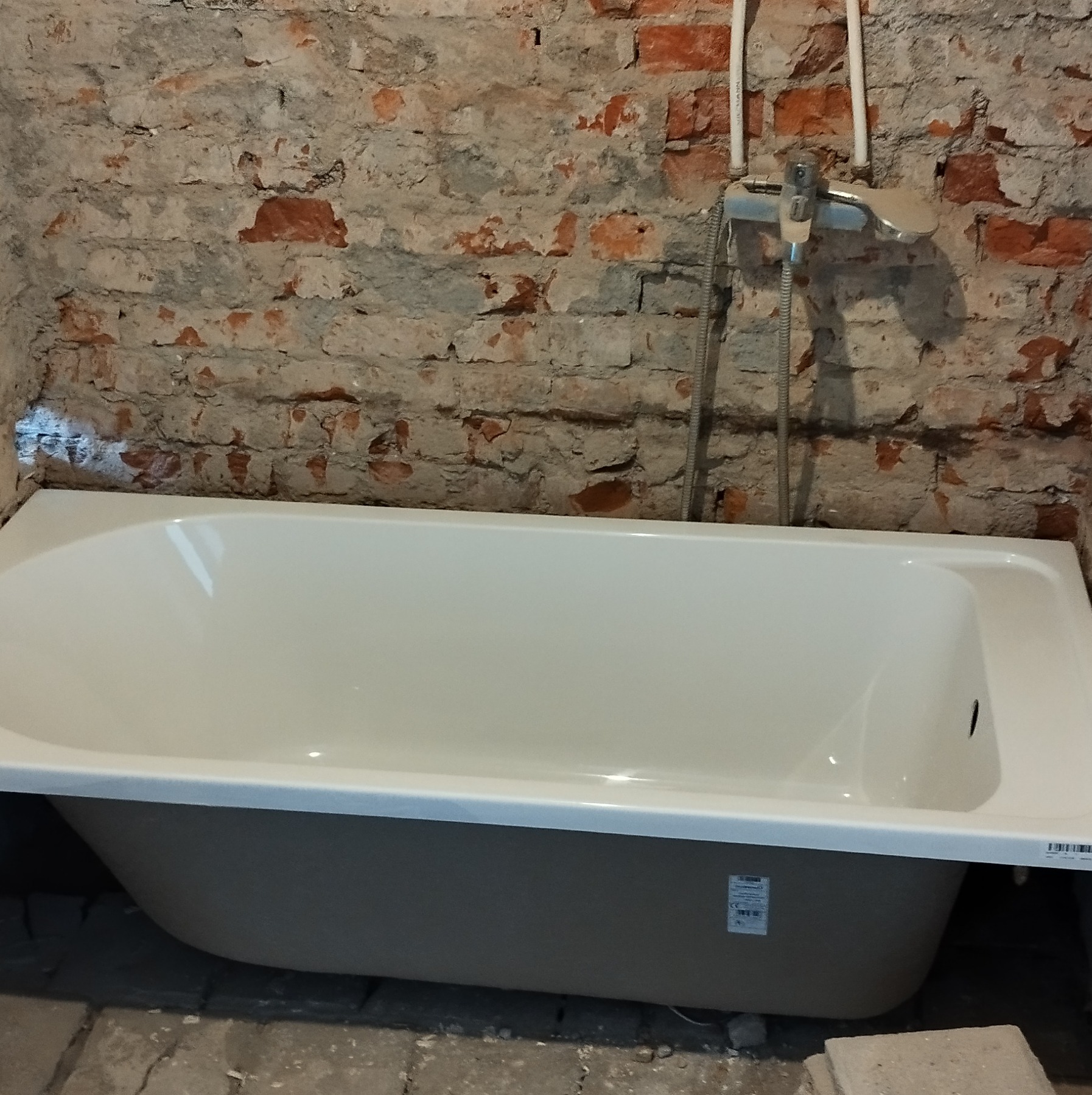 Nowa, biała wanna akrylowa z szarą obudową, częściowo osadzona w surowej łazience z odsłoniętą, ceglaną ścianą i widocznymi rurami oraz baterią prysznicową.