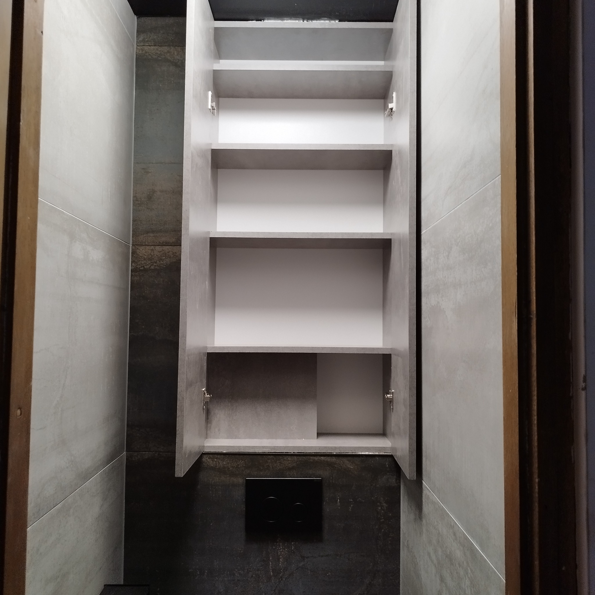 Otwarta szafka łazienkowa z trzema półkami, zamontowana nad panelem sterującym spłuczki, otoczona szarymi i ciemnymi płytkami imitującymi kamień.