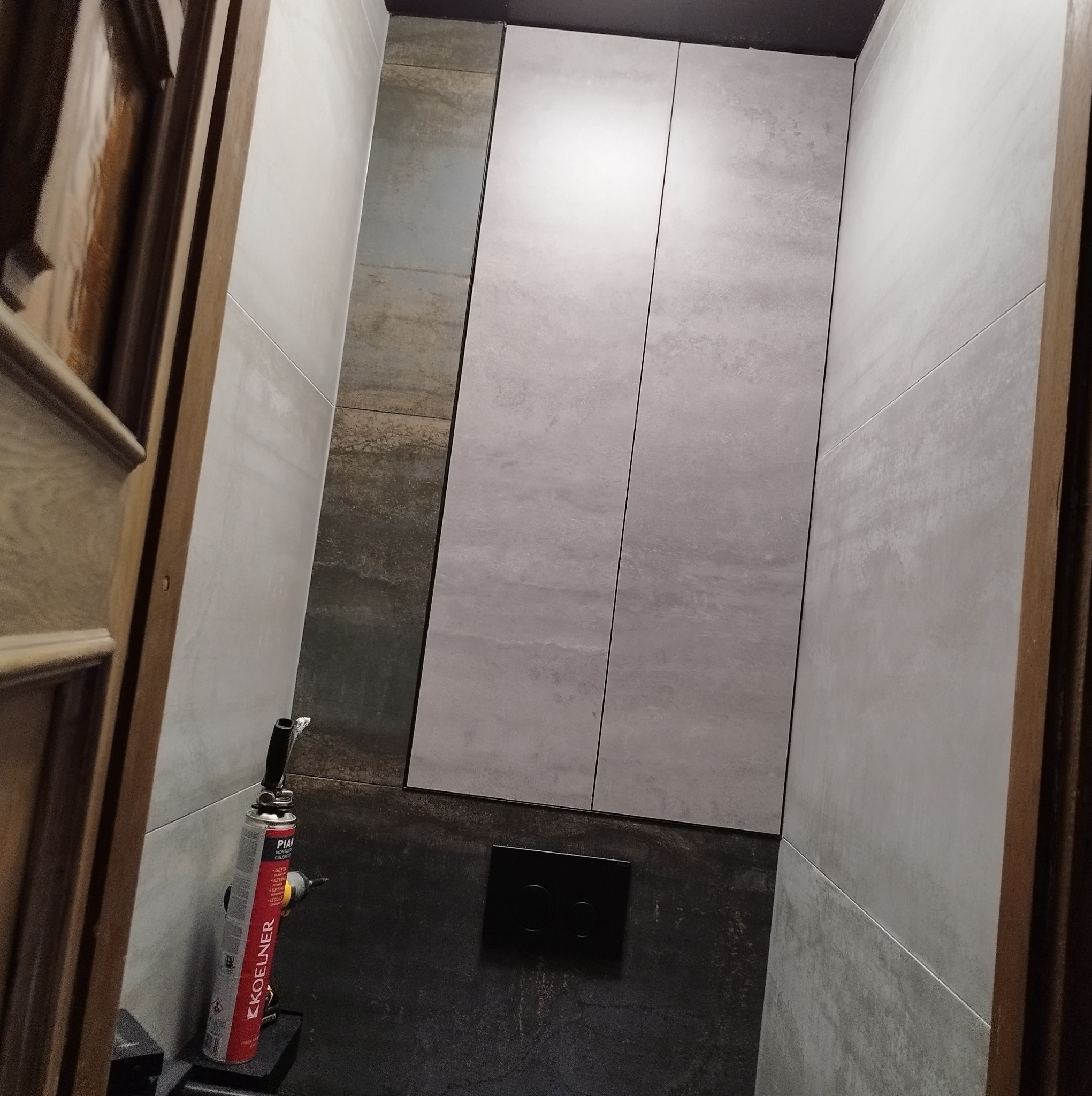 Wnętrze łazienki w trakcie prac wykończeniowych, widoczne szare płytki na ścianach, czarny panel spłuczki oraz pistolet do pianki montażowej Koelner stojący na podłodze.