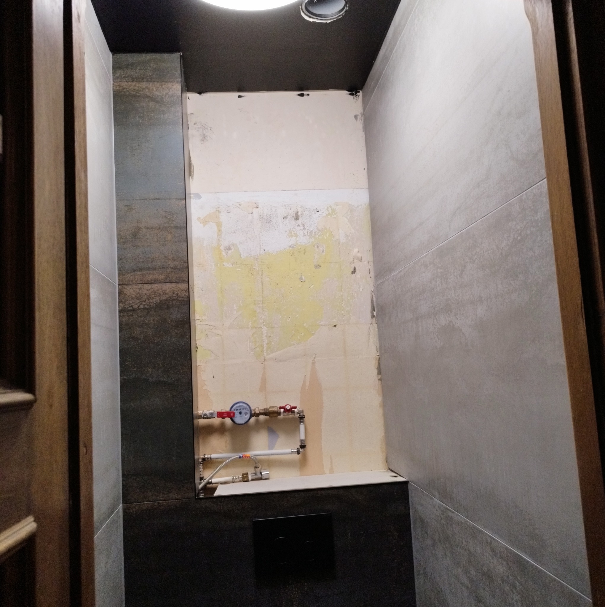 Łazienka w trakcie remontu: częściowo położone szare płytki imitujące beton, fragment ściany z resztkami starej glazury, widoczne rury i zawory wodne, czarny panel sterujący spłuczką.