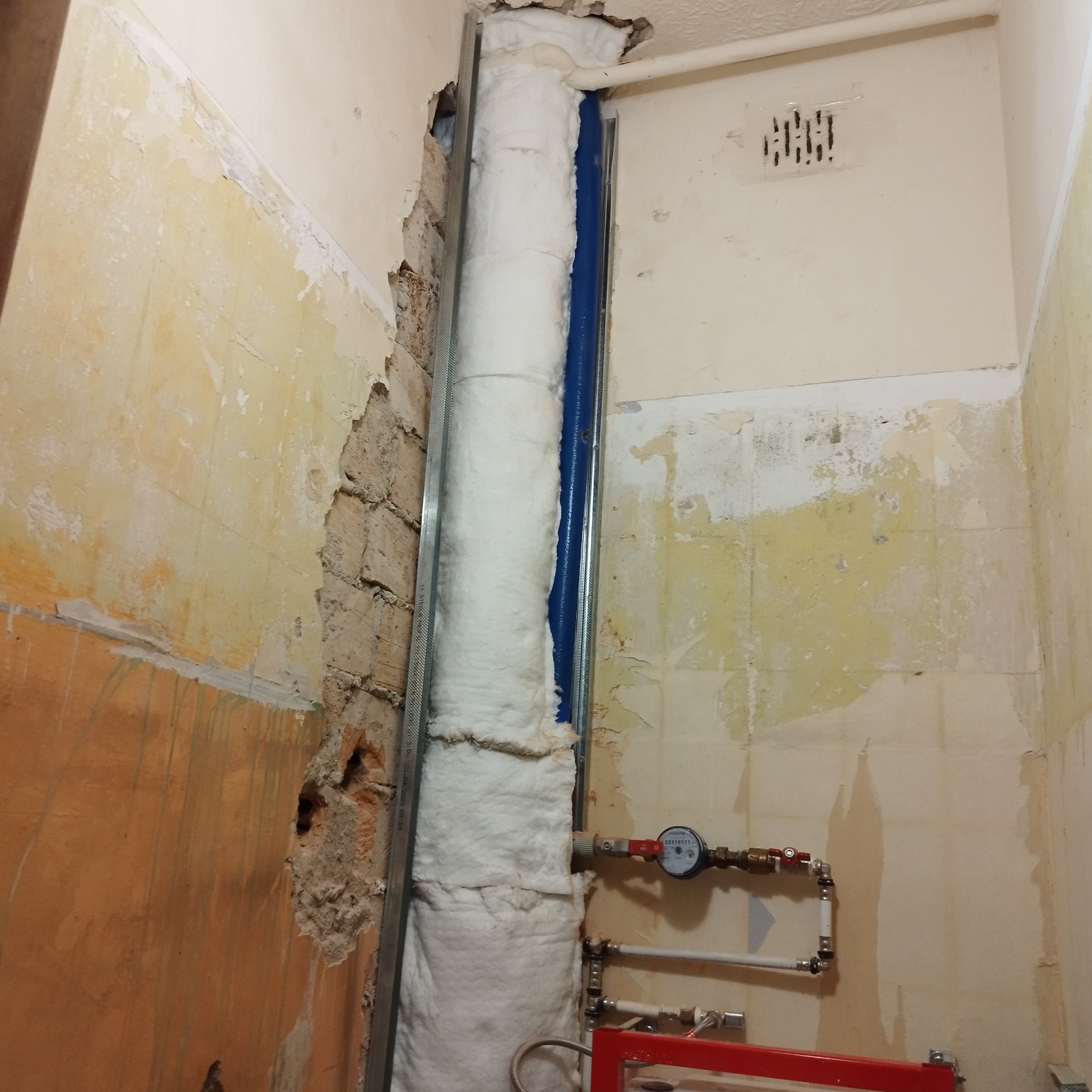 Odsłonięta ściana z instalacją wodną i rurą owiniętą izolacją, zabezpieczona metalowym profilem, widoczne ślady po usuniętych płytkach i starej farbie.