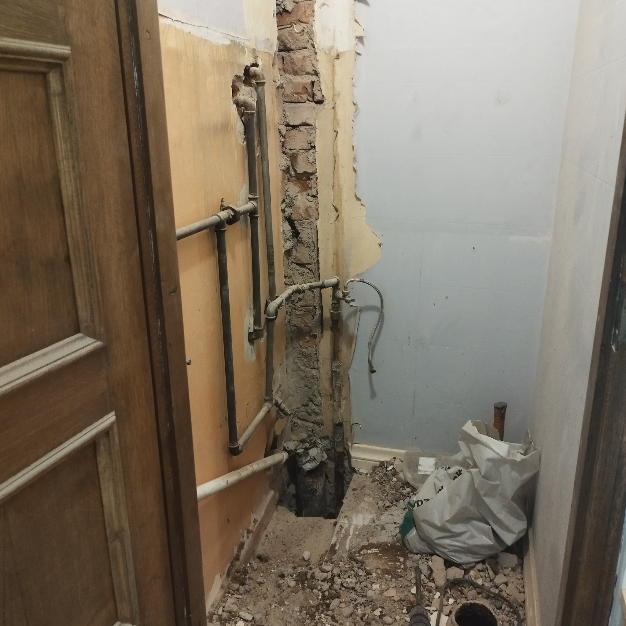 Łazienka w trakcie remontu: odsłonięte rury wodno-kanalizacyjne w ścianie, widoczny fragment muru z cegły, gruz na podłodze, częściowo zerwane płytki, otwarta przestrzeń pod podłogą.