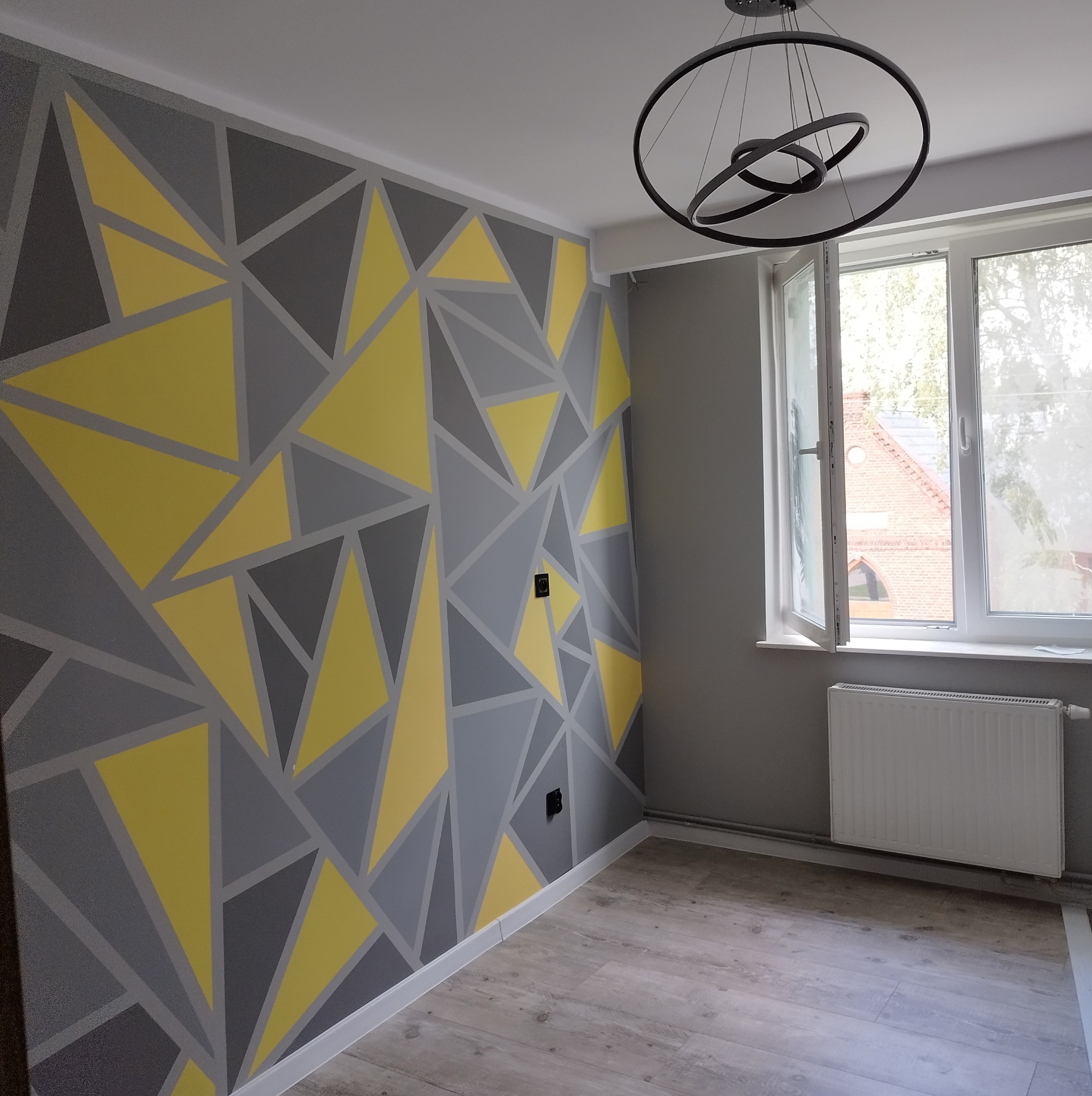 Pokój po remoncie z szaro-żółtą geometryczną dekoracją malarską na ścianie, nowoczesna lampa wisząca i otwarte okno z widokiem na ceglany budynek.