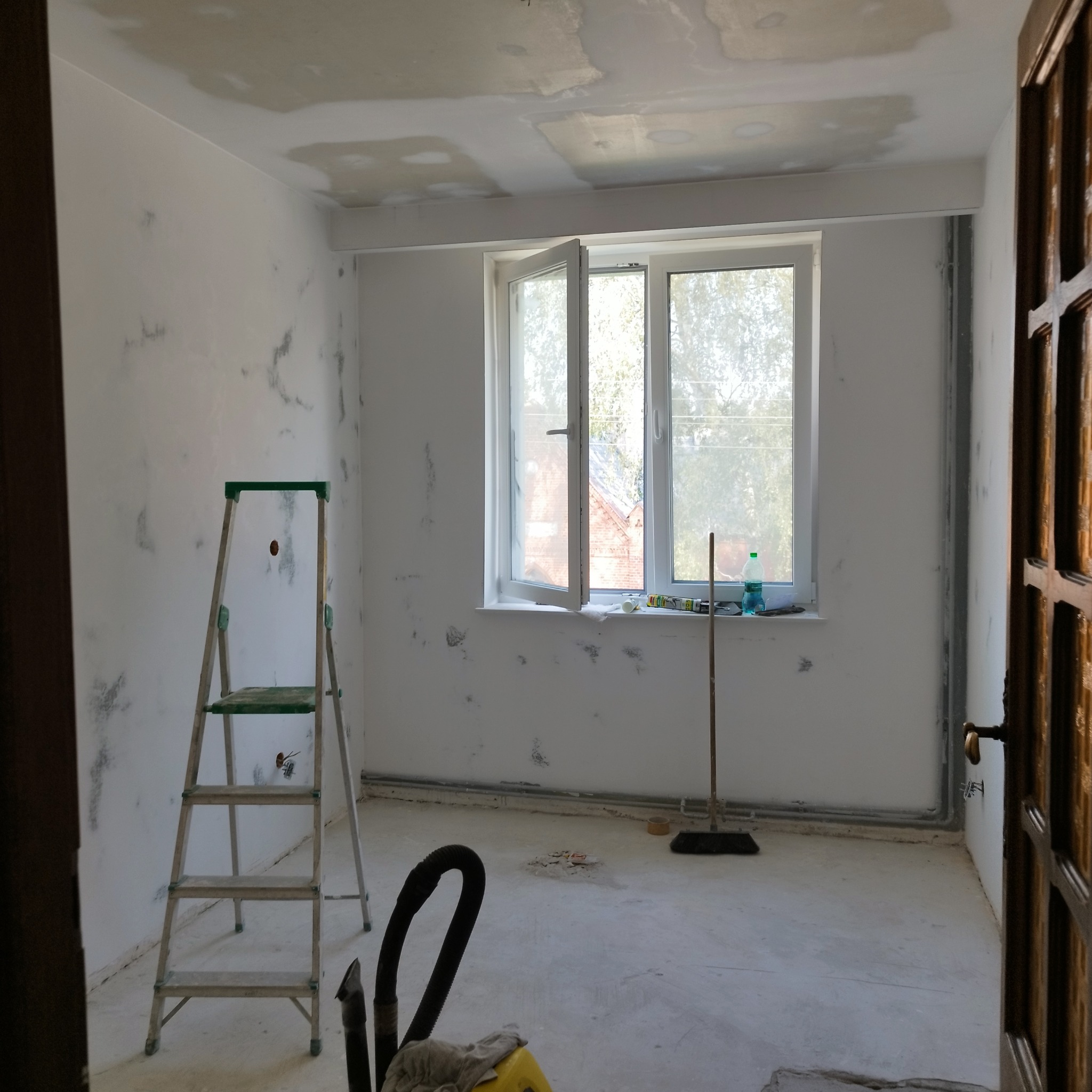 Pokój w trakcie przygotowania do malowania: ściany z widocznymi śladami po szpachlowaniu, drabina, otwarte okno z widokiem na zewnątrz, narzędzia malarskie i akcesoria na parapecie, oraz odkurzacz...