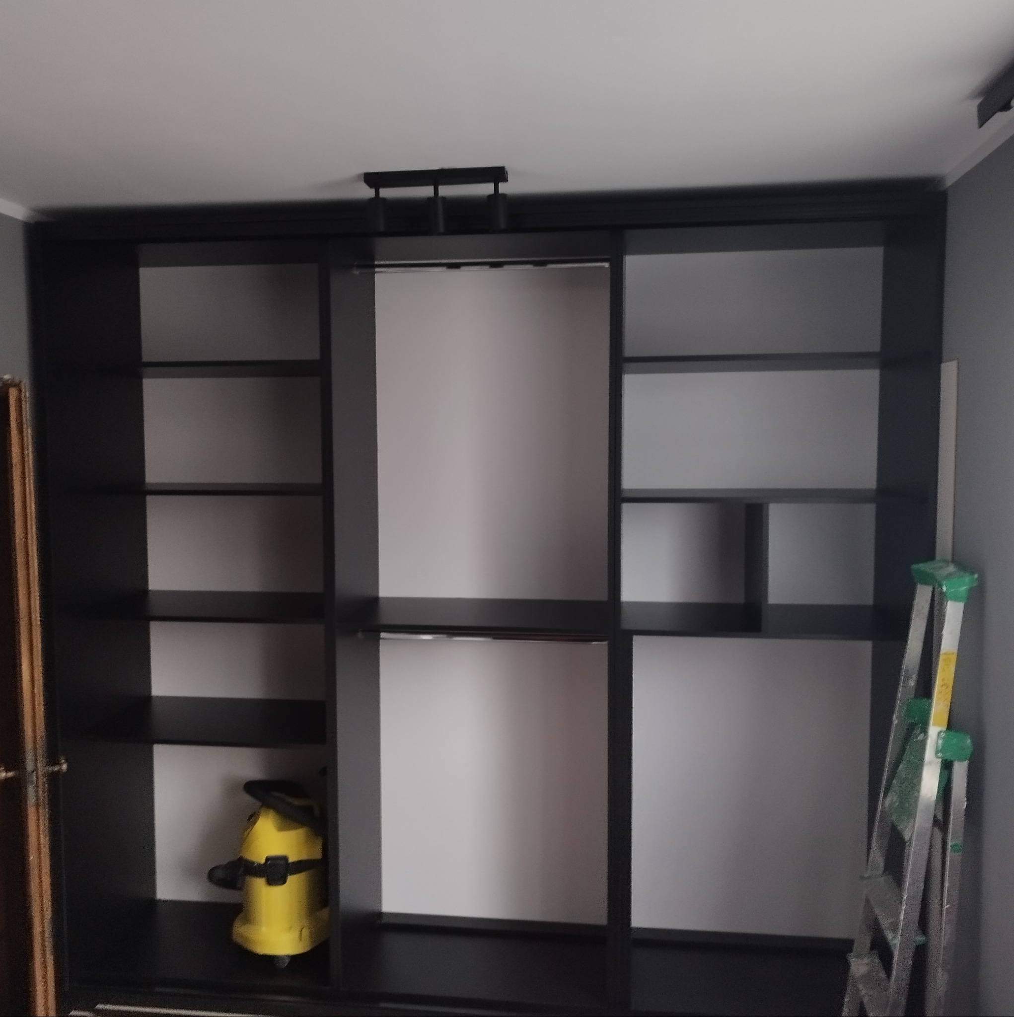 Czarna szafa wnękowa z przesuwnymi drzwiami, częściowo pusta, z żółtym odkurzaczem przemysłowym w jednej z półek i aluminiową drabiną opartą o ścianę po prawej stronie.