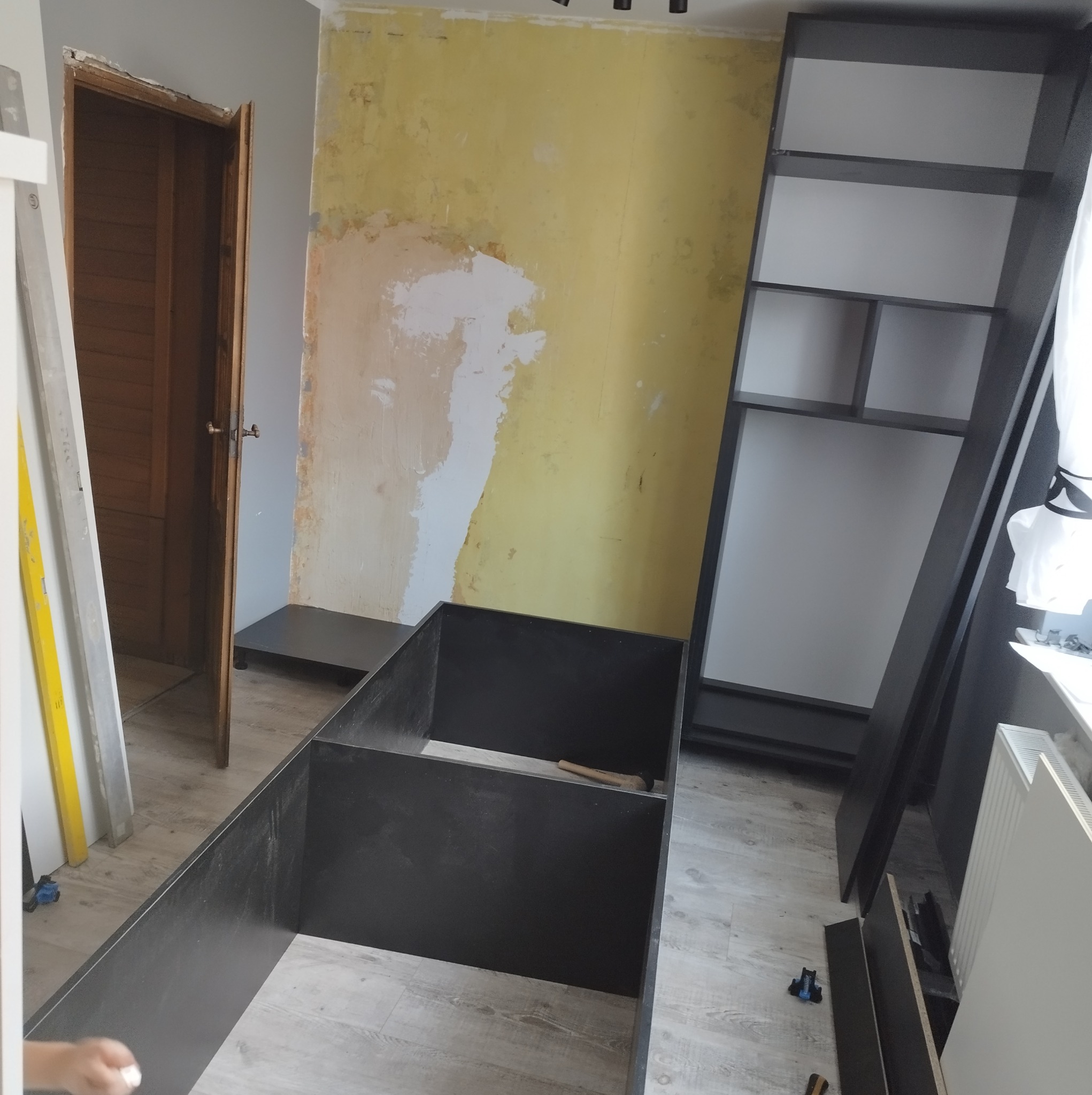 Częściowo zmontowany, czarny regał z białymi półkami, ustawiony w pokoju z żółtą ścianą w trakcie remontu, widoczna otwarta drewniana rama drzwi i poziomica.