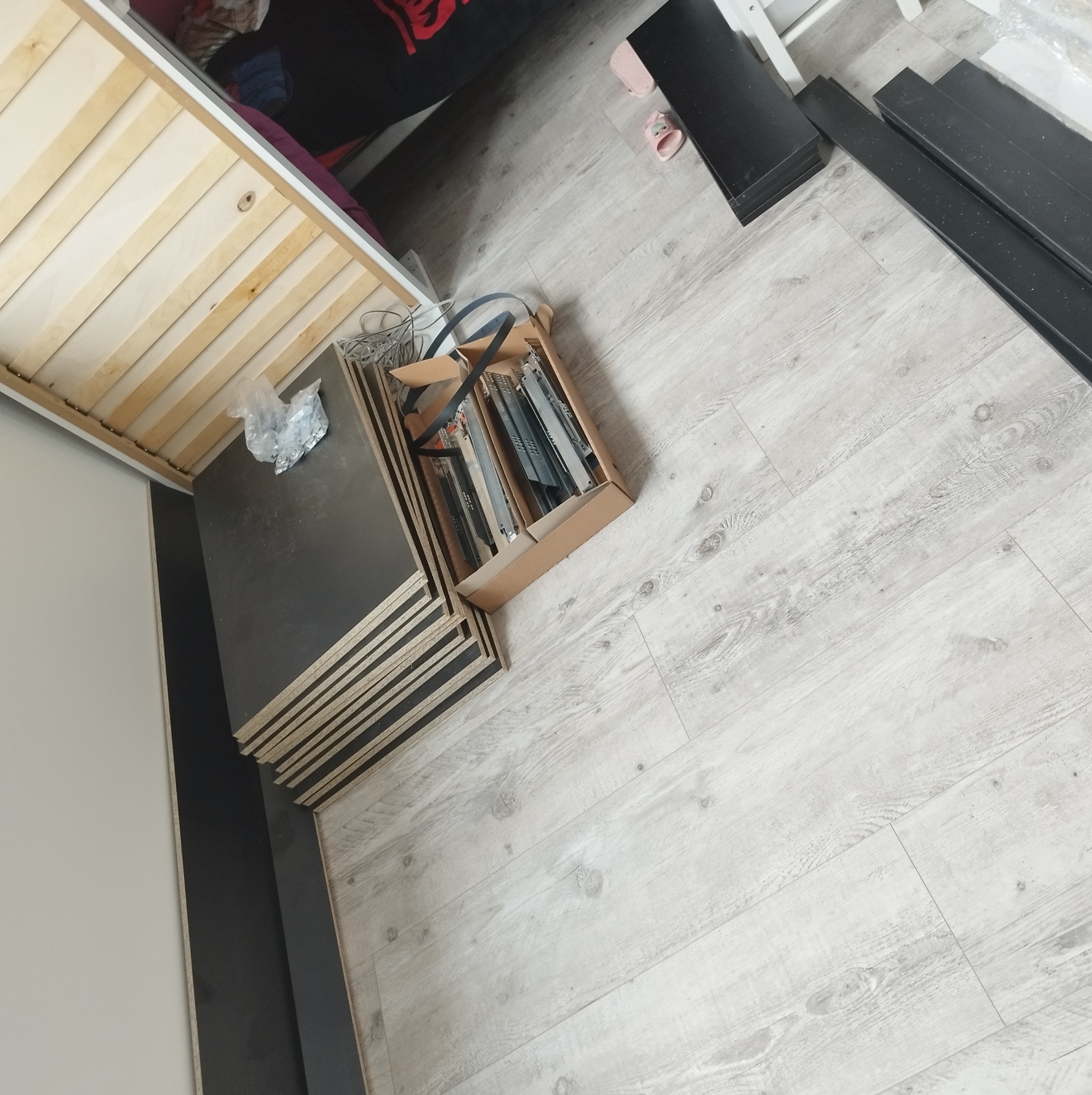 Przygotowane elementy do montażu mebli: płyty, prowadnice do szuflad w kartonie, widoczne na jasnej podłodze z paneli.