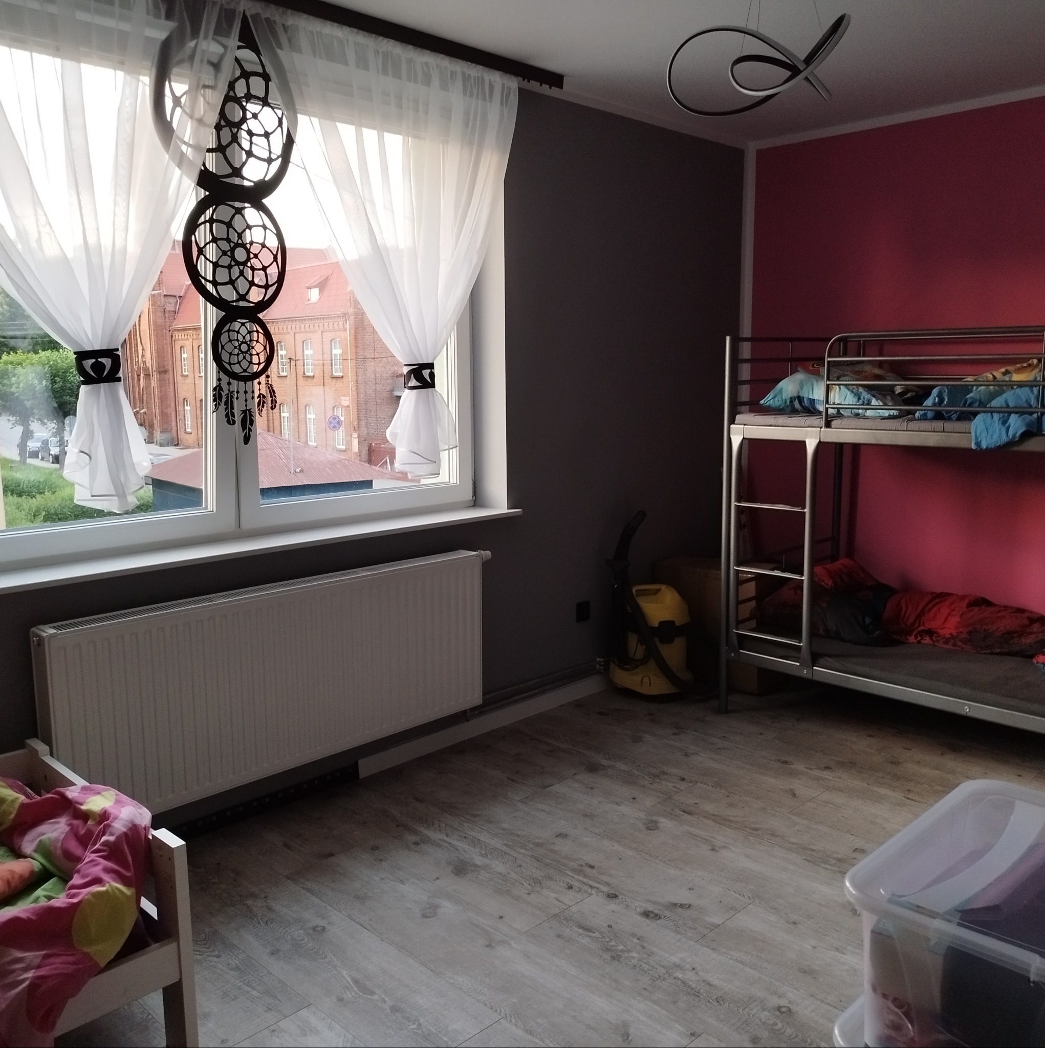Pokój dziecięcy z łóżkiem piętrowym, ścianą w kolorze fuksji i szarą, drewnianą podłogą. Na oknie białe zasłony z czarnymi ozdobami i łapacz snów.
