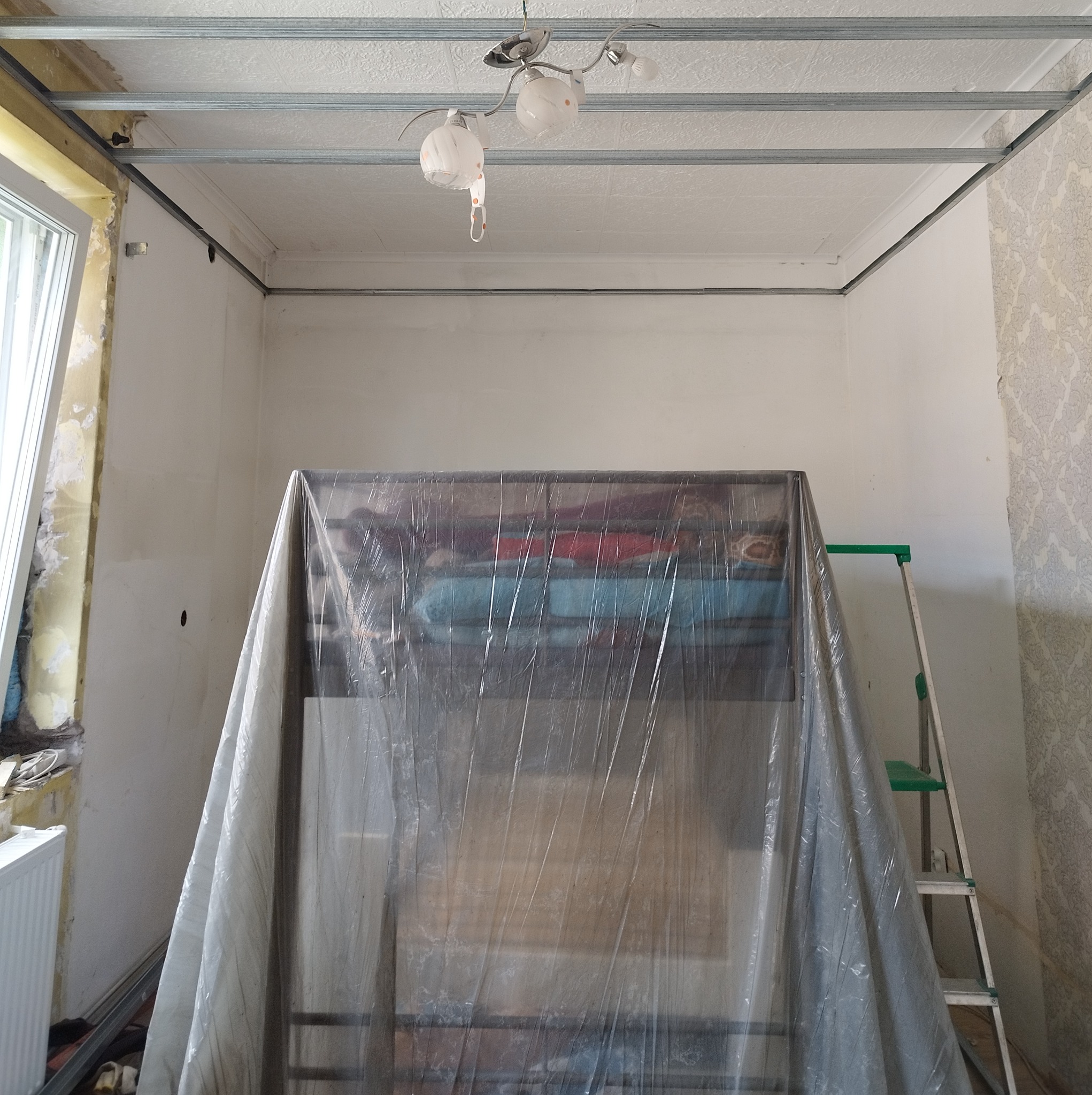 Pokój w trakcie remontu, meble zabezpieczone folią malarską, widoczne profile sufitowe, ściany przygotowane do malowania, drabina oparta o ścianę.