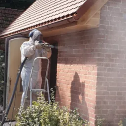 Pracownik w kombinezonie ochronnym i słuchawkach, stojąc na drabinie, czyści ceglaną ścianę budynku za pomocą specjalistycznego sprzętu. Widoczny efekt czyszczenia na ścianie.