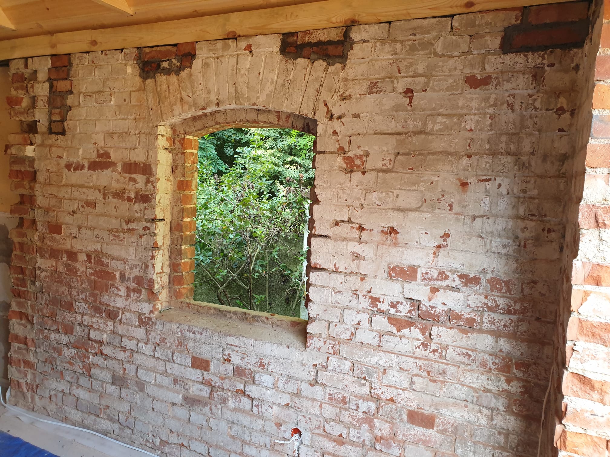 Ceglana ściana z oknem o łukowym kształcie, częściowo pokryta białą farbą, z widocznymi śladami po usuniętej powłoce. Przez okno widać zieleń drzew.