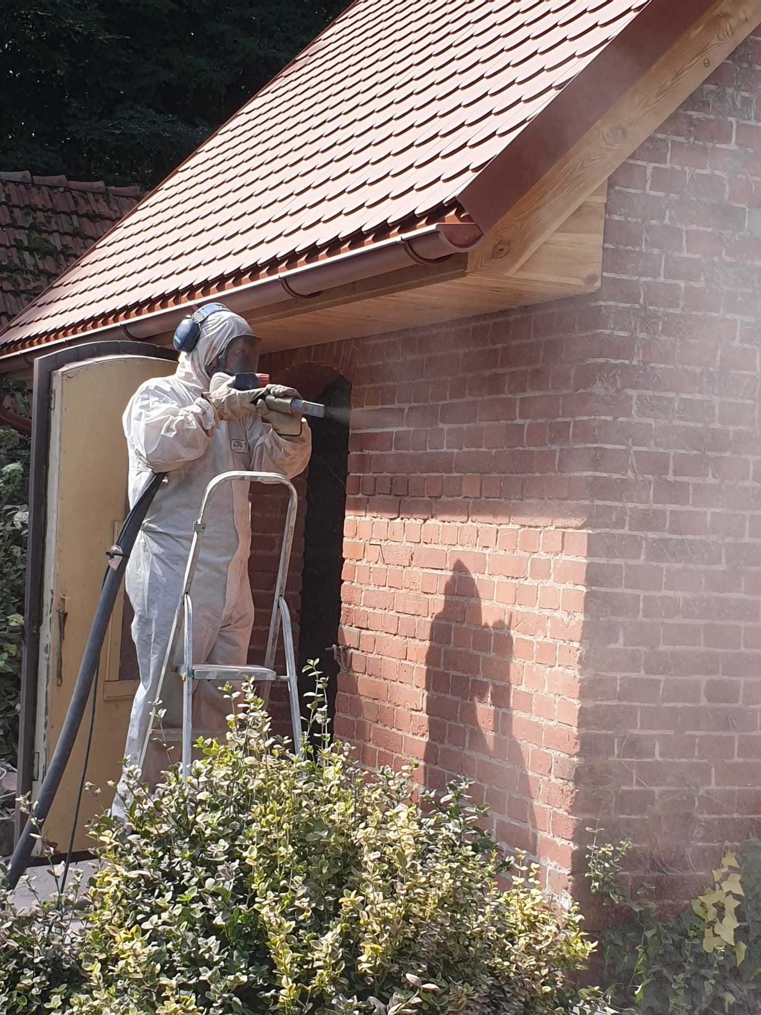 Pracownik w kombinezonie ochronnym i słuchawkach, stojąc na drabinie, czyści ceglaną ścianę budynku za pomocą specjalistycznego sprzętu. Widoczny efekt czyszczenia na ścianie.