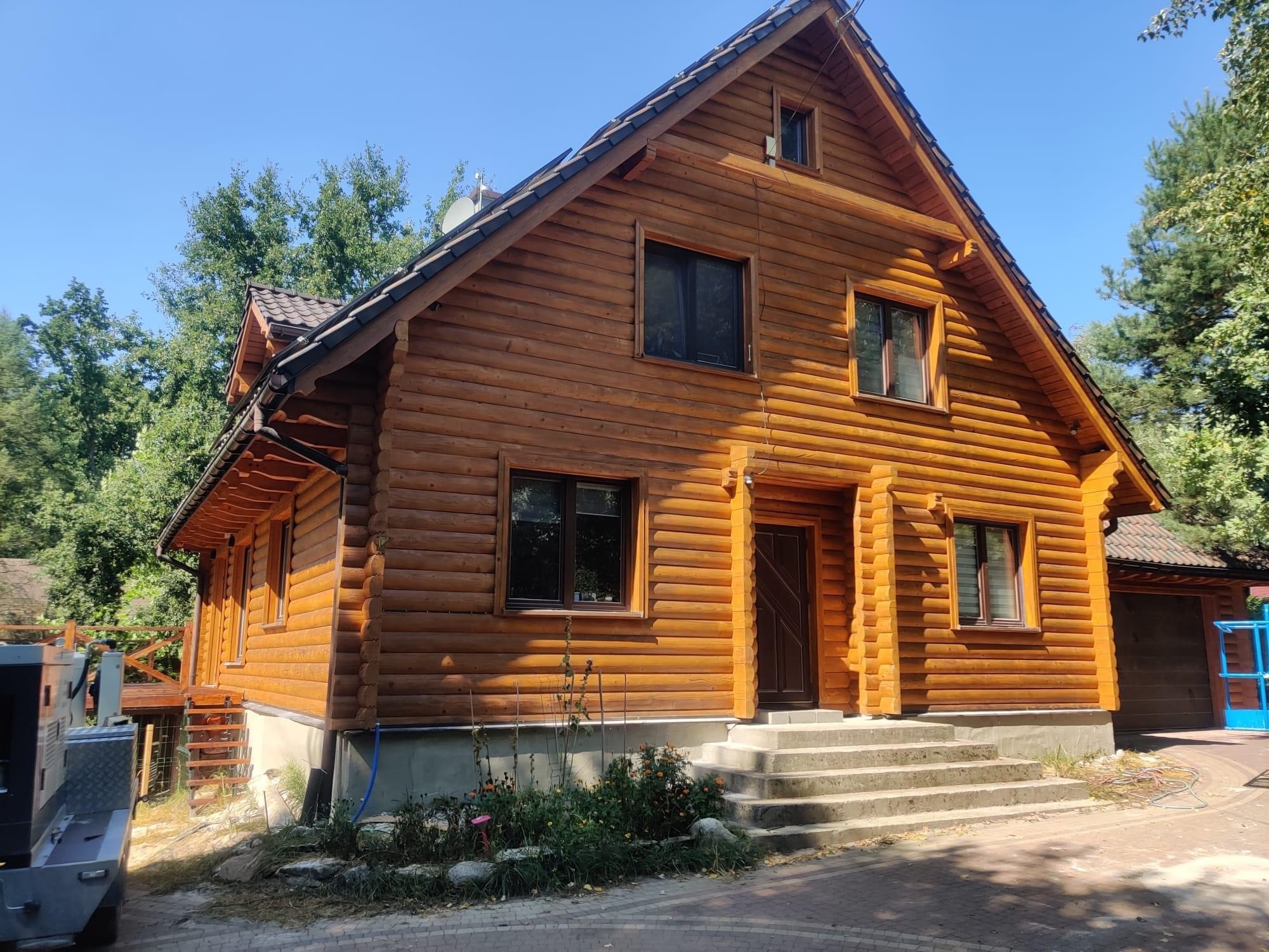 Dwukondygnacyjny dom z bali drewnianych z ciemnymi oknami i drzwiami, widoczny z poziomu chodnika z kostki brukowej w słoneczny dzień.