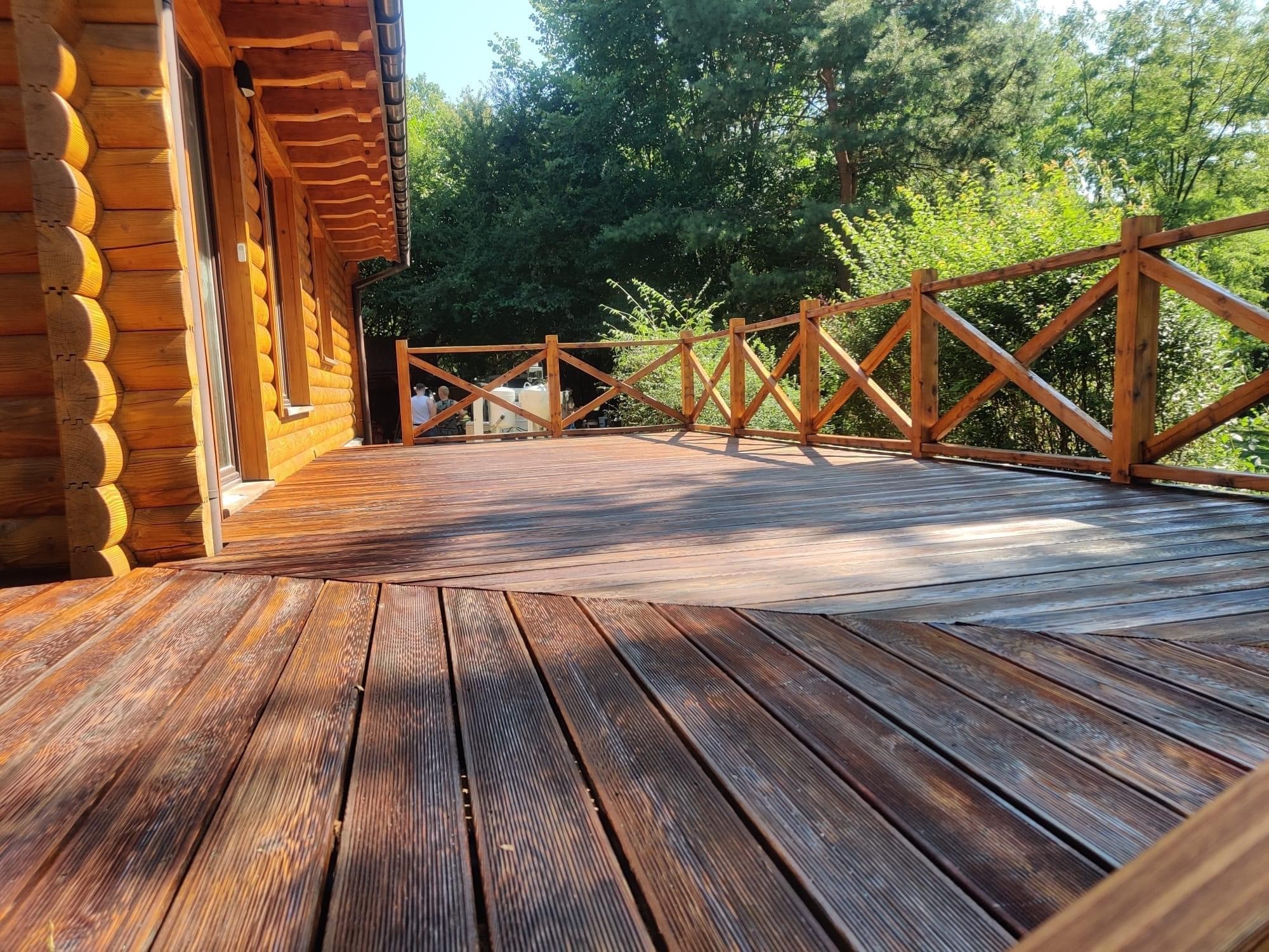 Drewniany taras przy domu z bali, zabezpieczony impregnatem, z dekoracyjną balustradą w stylu rustykalnym, otoczony zielenią.