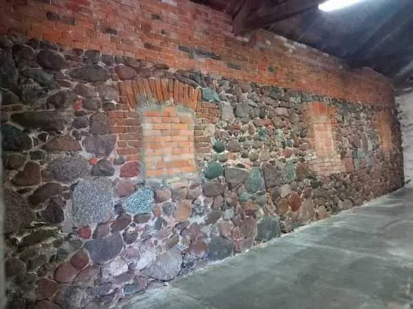Fragment ściany z kamienia polnego i cegły, z zamurowanymi otworami okiennymi, widoczne ślady wcześniejszych napraw i renowacji.