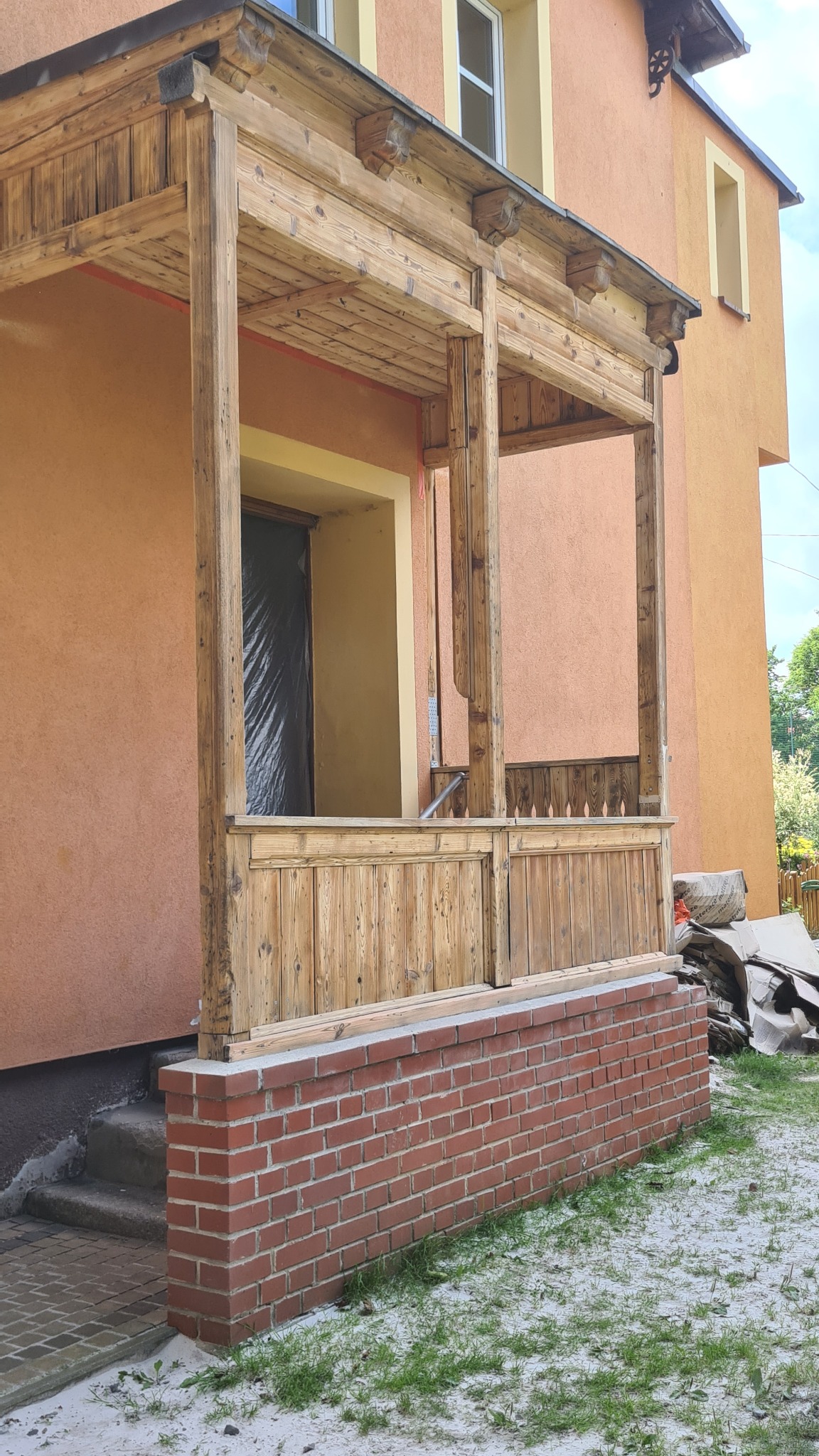 Drewniany ganek z ceglaną podmurówką w trakcie renowacji, zabezpieczone wejście folią, widoczne ślady obróbki drewna.