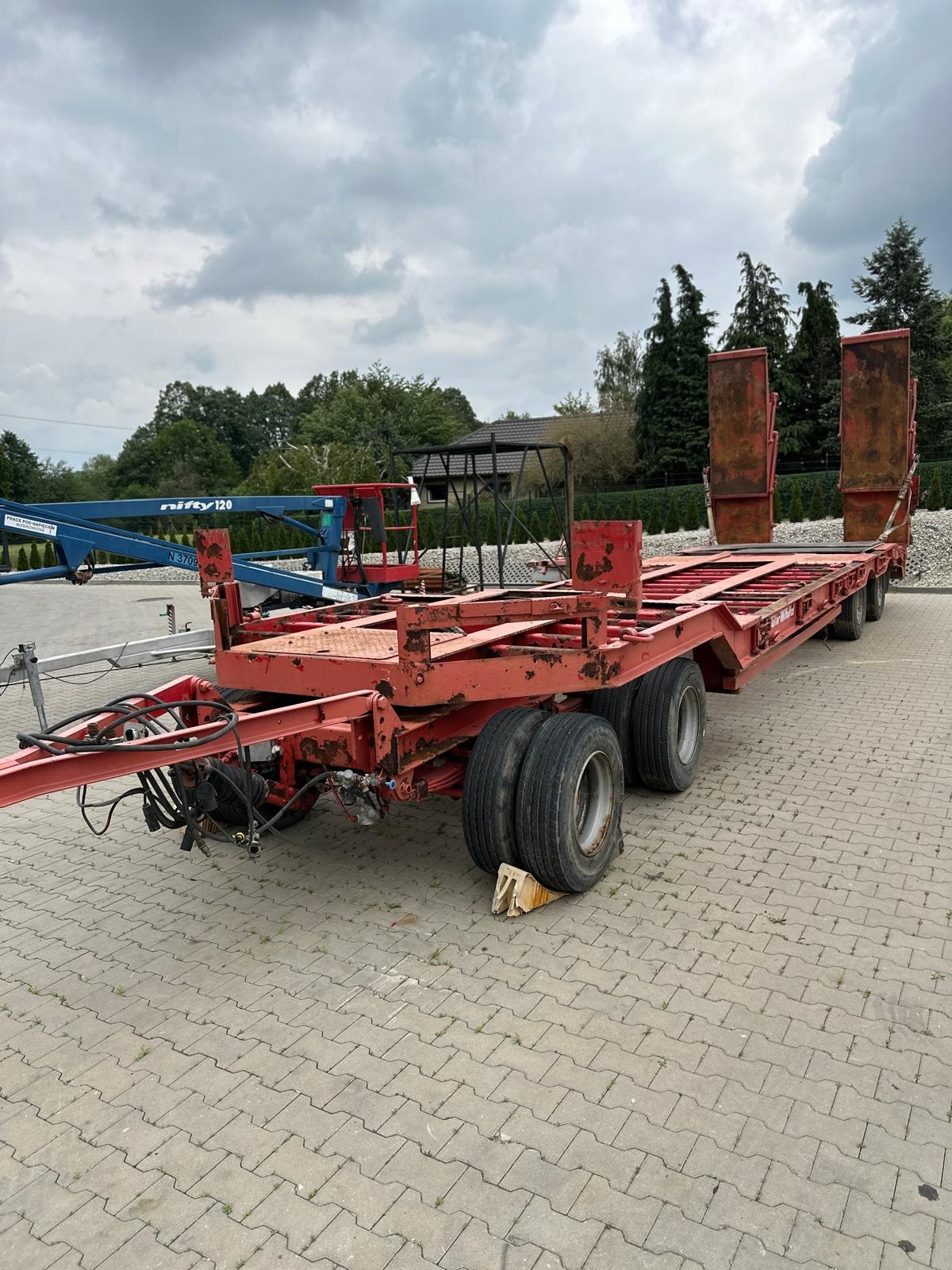 Czerwona, używana platforma transportowa z czterema osiami, stojąca na kostce brukowej, widoczna z boku, z elementami korozji i zużycia, z podłożonym klinem pod kołem, w tle widoczny podnośnik...