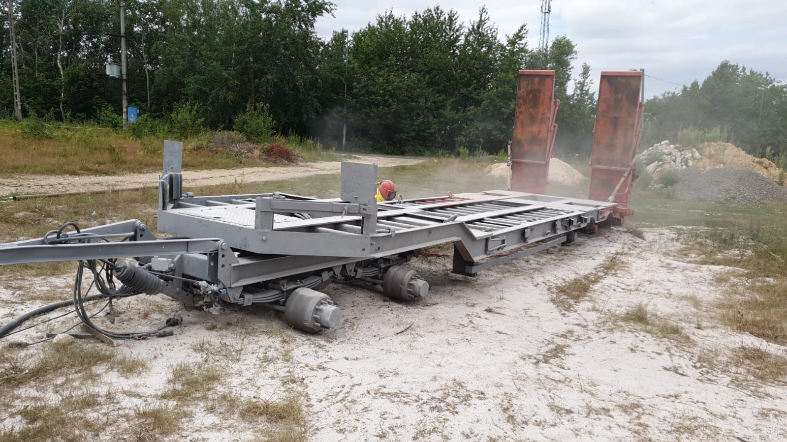 Przyczepa niskopodwoziowa po piaskowaniu, widoczne ślady pyłu, czerwony kask na platformie, tło z drzewami i hałdami piasku.