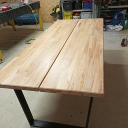 ADM Usługi Spawalnicze - Blat stołu z jasnego drewna klejonego, złożony z dwóch części, na czarnych, metalowych nogach w warsztacie.