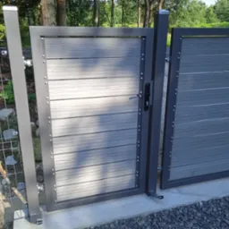 Nowoczesna brama wjazdowa z szarego metalu i paneli imitujących drewno, widoczna z bliska, z fragmentem ogrodzenia siatkowego w tle.