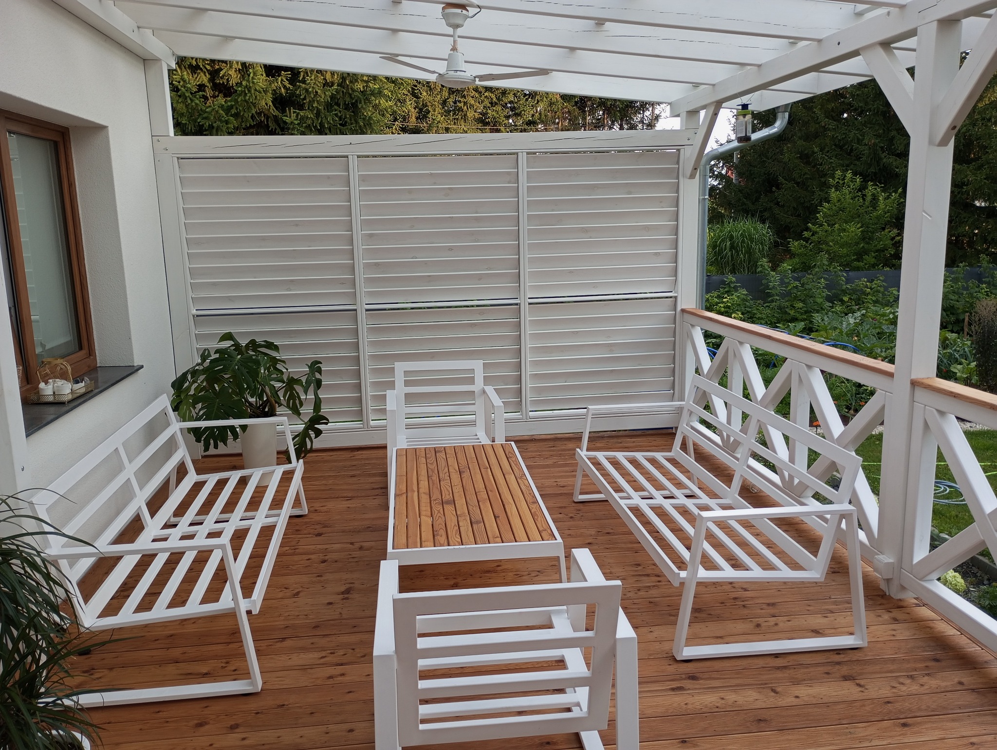 Zadaszony taras z drewnianą podłogą, białymi meblami ogrodowymi o konstrukcji metalowej, przesłonięty ażurową ścianą i pergolą, z widocznymi roślinami w tle.