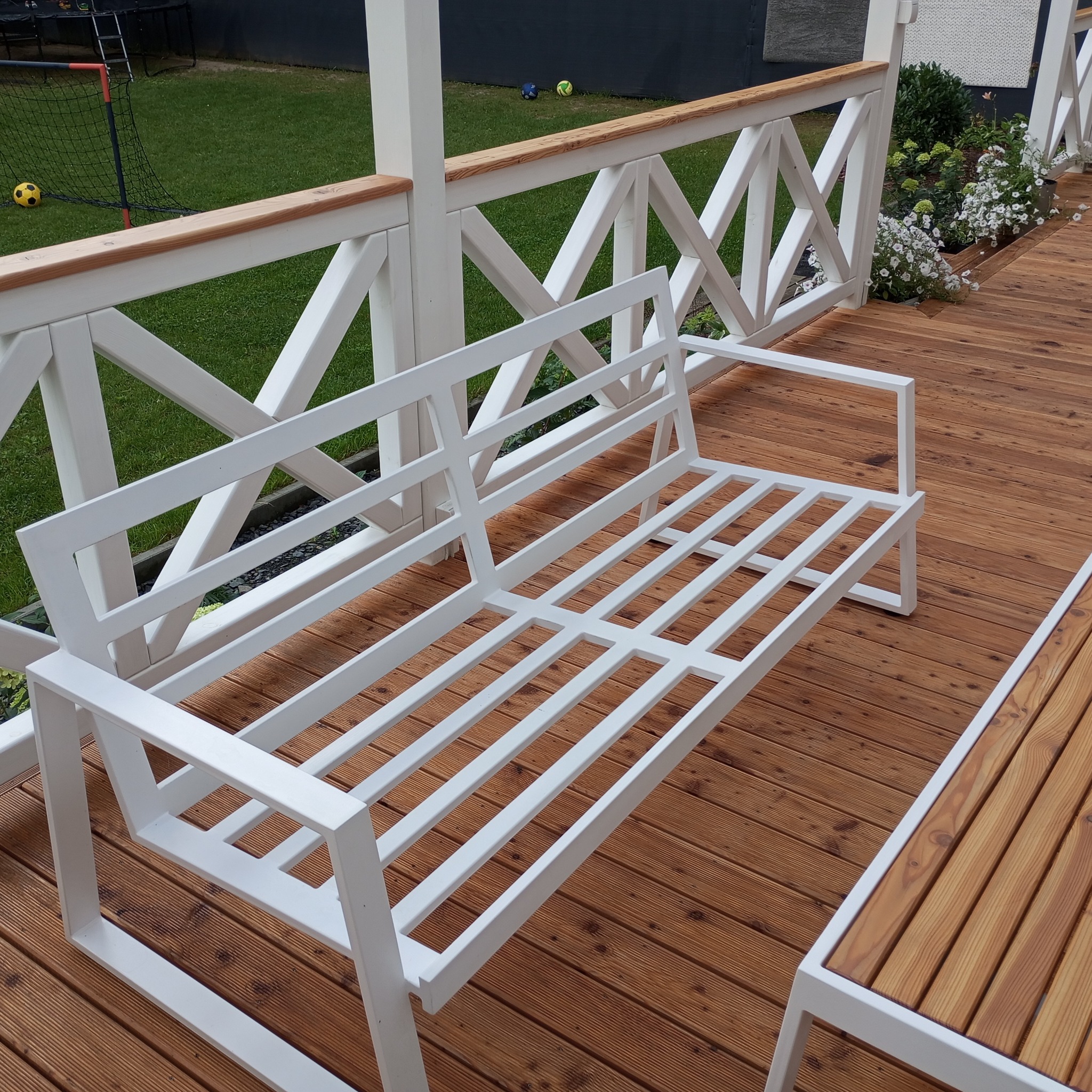 Biała, metalowa ławka o minimalistycznym designie na drewnianym tarasie, z widoczną balustradą z białymi słupkami i drewnianą poręczą, w tle trawnik i elementy ogrodu.