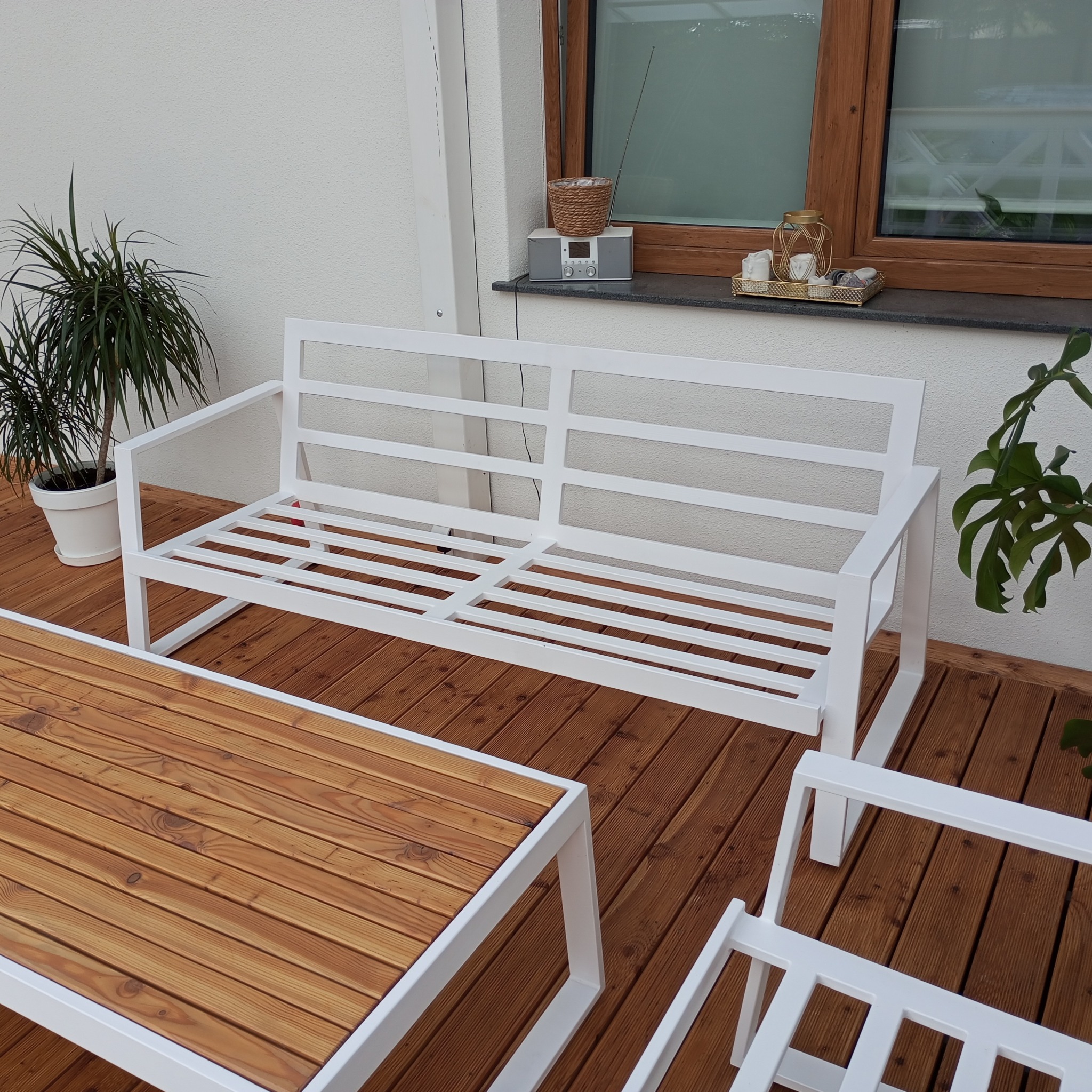 Biały metalowy zestaw mebli ogrodowych: sofa i stolik z drewnianym blatem, na drewnianym tarasie, z dekoracjami na parapecie okiennym i roślinami doniczkowymi.