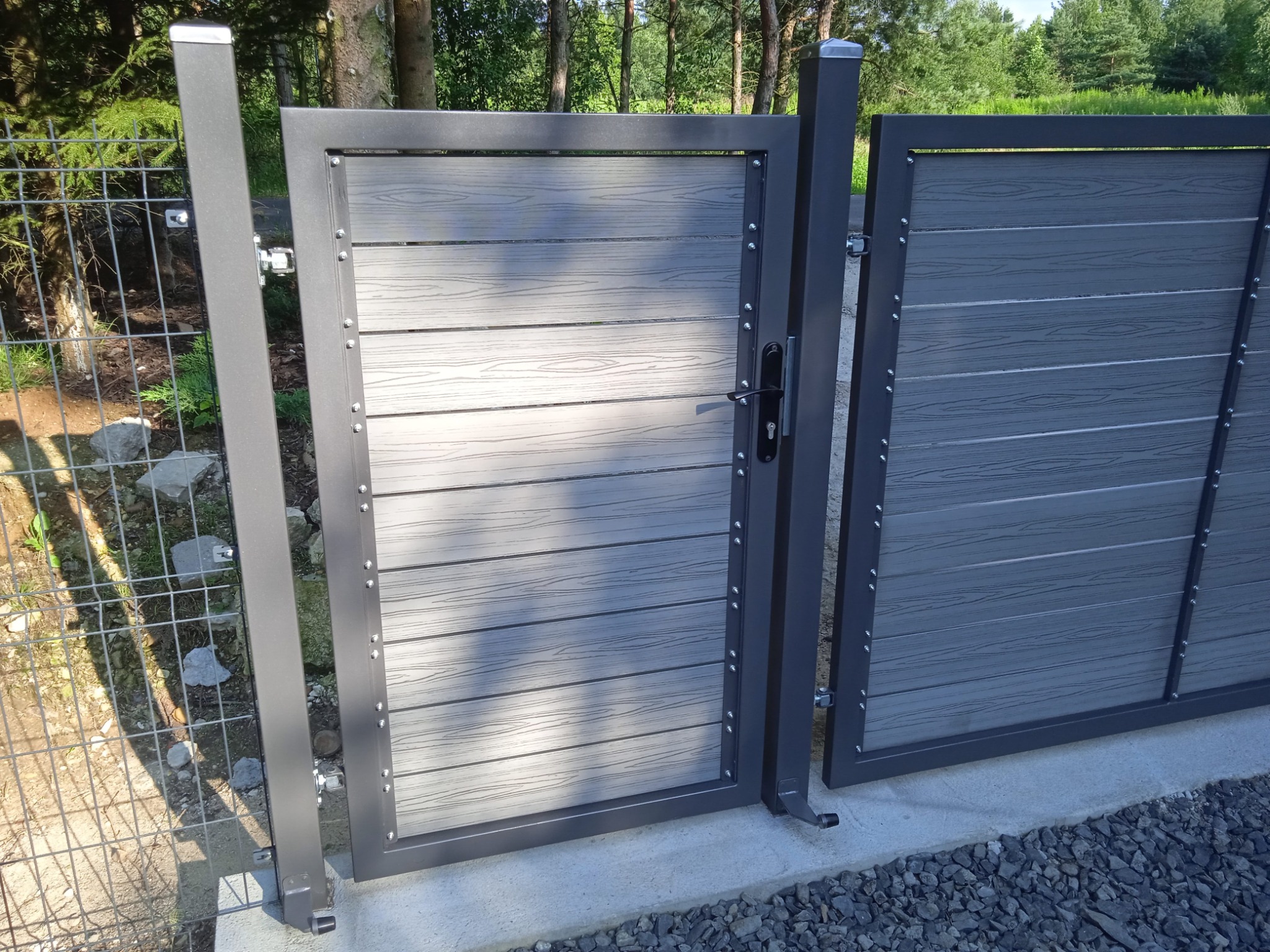Nowoczesna brama wjazdowa z szarego metalu i paneli imitujących drewno, widoczna z bliska, z fragmentem ogrodzenia siatkowego w tle.