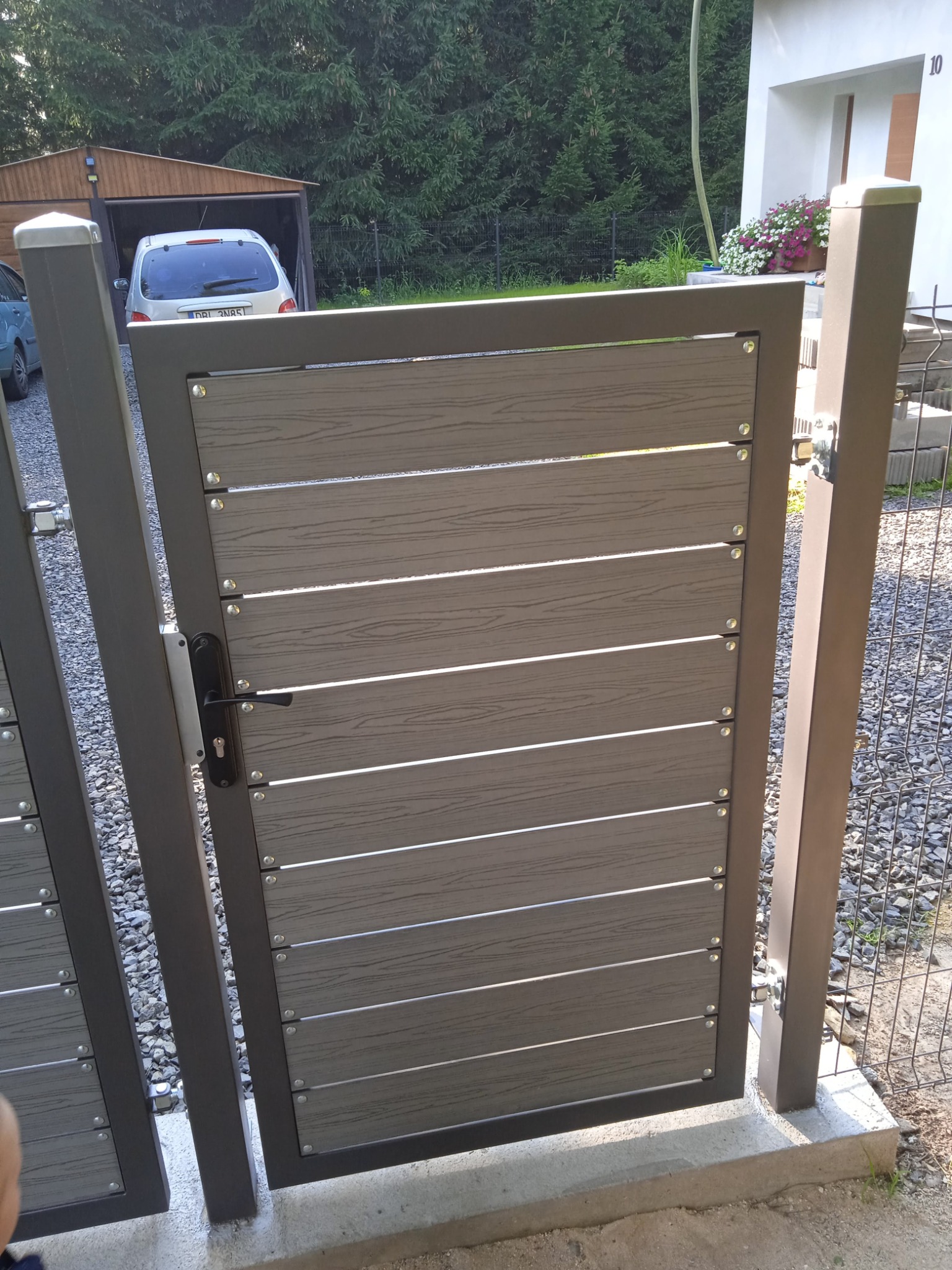 Nowoczesna brama wjazdowa z poziomych paneli imitujących drewno, z metalową ramą w kolorze grafitowym, zamontowana na posesji z widocznym domem i garażem w tle.