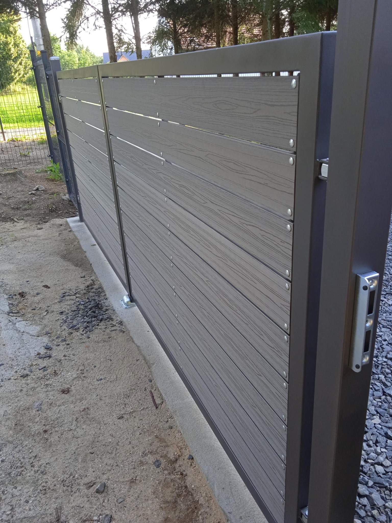 Nowoczesna brama wjazdowa przesuwna z poziomymi panelami imitującymi drewno, zamontowana na betonowej podstawie, widok z boku na mechanizm zamykający.