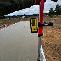 Kalvia Radosław Gazarkiewicz - Pomiar wylewki betonowej z użyciem niwelatora laserowego Nivel na łacie, widoczne rękawice robocze i fragment terenu budowy.