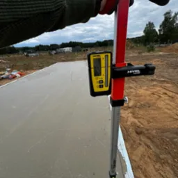 Pomiar wylewki betonowej z użyciem niwelatora laserowego Nivel na łacie, widoczne rękawice robocze i fragment terenu budowy.