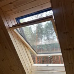 Wnętrze domu szkieletowego z drewnianą boazerią i oknem dachowym uchylonym do góry, widok na las sosnowy.