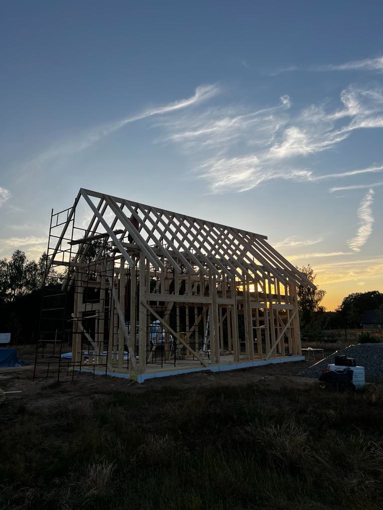 Wznoszenie konstrukcji szkieletowej domu jednorodzinnego o zachodzie słońca, widoczny częściowo ukończony szkielet z drewnianych belek i rusztowania.