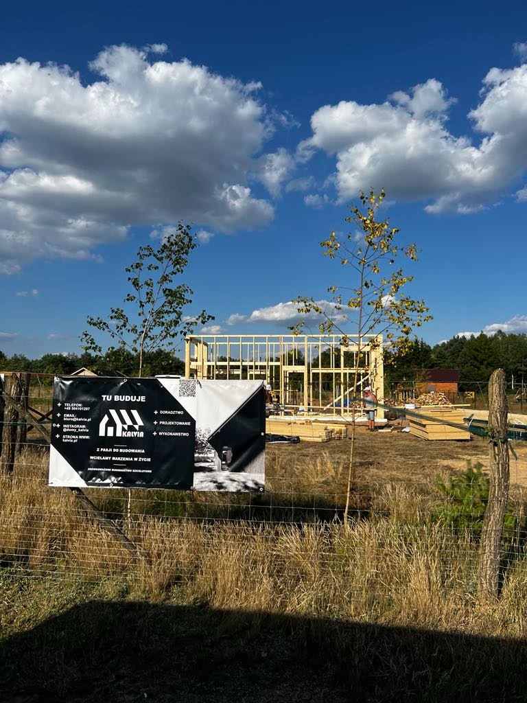 Wznoszenie konstrukcji szkieletowej domu na działce, widoczny baner reklamowy firmy budowlanej, błękitne niebo z obłokami w tle.