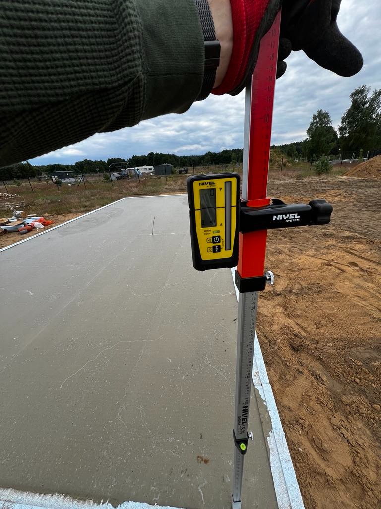 Pomiar wylewki betonowej z użyciem niwelatora laserowego Nivel na łacie, widoczne rękawice robocze i fragment terenu budowy.