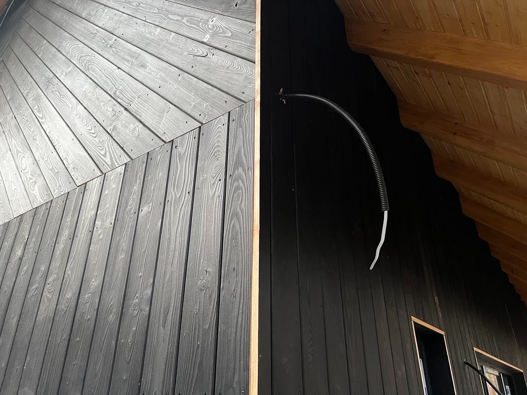 Fragment elewacji budynku z ciemnego drewna, z widocznym fragmentem dachu i instalacją elektryczną w peszlu, ukazujący detale konstrukcyjne.