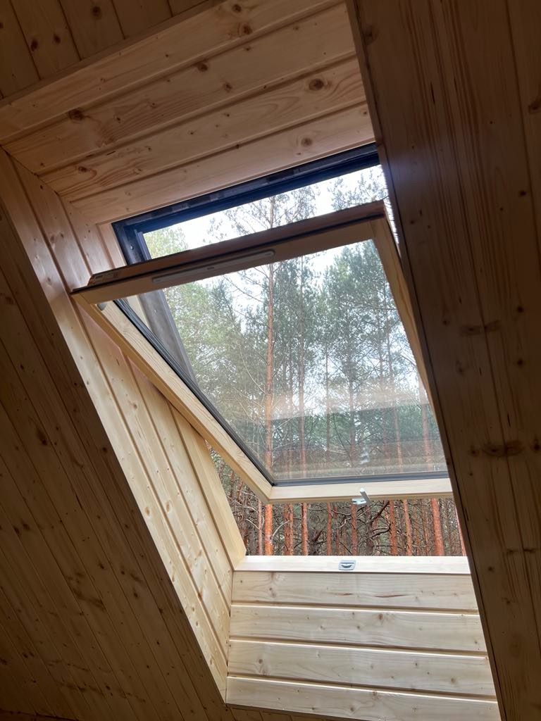 Wnętrze domu szkieletowego z drewnianą boazerią i oknem dachowym uchylonym do góry, widok na las sosnowy.