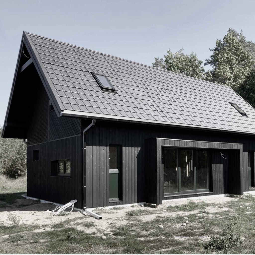 Nowoczesny, czarny dom z drewna z szarym dachem, widok z boku, z widocznymi oknami i rynnami, na tle drzew.