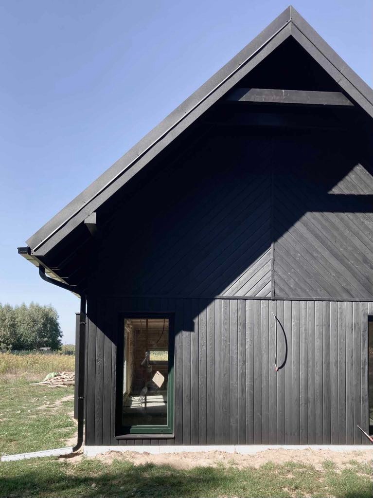 Ciemna, drewniana elewacja domu w trakcie budowy, widoczne okno bez wykończenia i niezagospodarowany teren wokół.