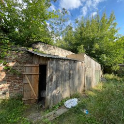 W001 SPÓŁKA Z OGRANICZONĄ ODPOWIEDZIALNOŚCIĄ - Stara, drewniana szopa z otwartymi drzwiami, obok ceglany budynek, zarośnięta trawa i śmieci na ziemi, widok w słoneczny dzień.