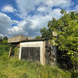 W001 SPÓŁKA Z OGRANICZONĄ ODPOWIEDZIALNOŚCIĄ - Stary, betonowy garaż z zardzewiałymi, metalowymi drzwiami, porośnięty trawą i krzewami, na tle błękitnego nieba z chmurami.