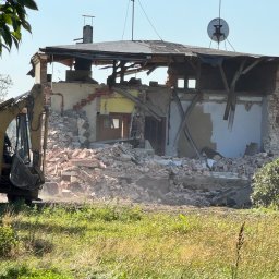 W001 SPÓŁKA Z OGRANICZONĄ ODPOWIEDZIALNOŚCIĄ - Żółta koparka częściowo widoczna z lewej strony, gruzowisko po rozbiórce budynku z widocznymi fragmentami ścian, okna i dachu, na dachu antena satelitarna, a w tle zielona łąka.