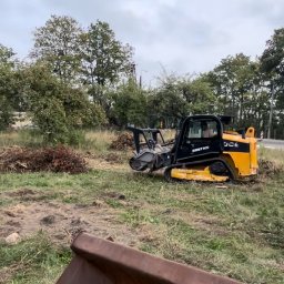 W001 SPÓŁKA Z OGRANICZONĄ ODPOWIEDZIALNOŚCIĄ - Żółto-czarna frezarka do pni drzew w trakcie pracy na zarośniętym terenie, widoczne ślady frezowania, w tle drzewa i droga.
