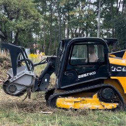 W001 SPÓŁKA Z OGRANICZONĄ ODPOWIEDZIALNOŚCIĄ - Żółto-czarna maszyna do frezowania pni drzew JCB 225T eco, z operatorem w kabinie, na zarośniętym terenie w tle drzewa.