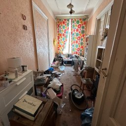 W001 SPÓŁKA Z OGRANICZONĄ ODPOWIEDZIALNOŚCIĄ - Długi, wąski korytarz zagracony meblami, kartonami, workami i sprzętami AGD, z kolorowymi zasłonami w tle, ukazujący stan pomieszczenia przed rozpoczęciem porządkowania.