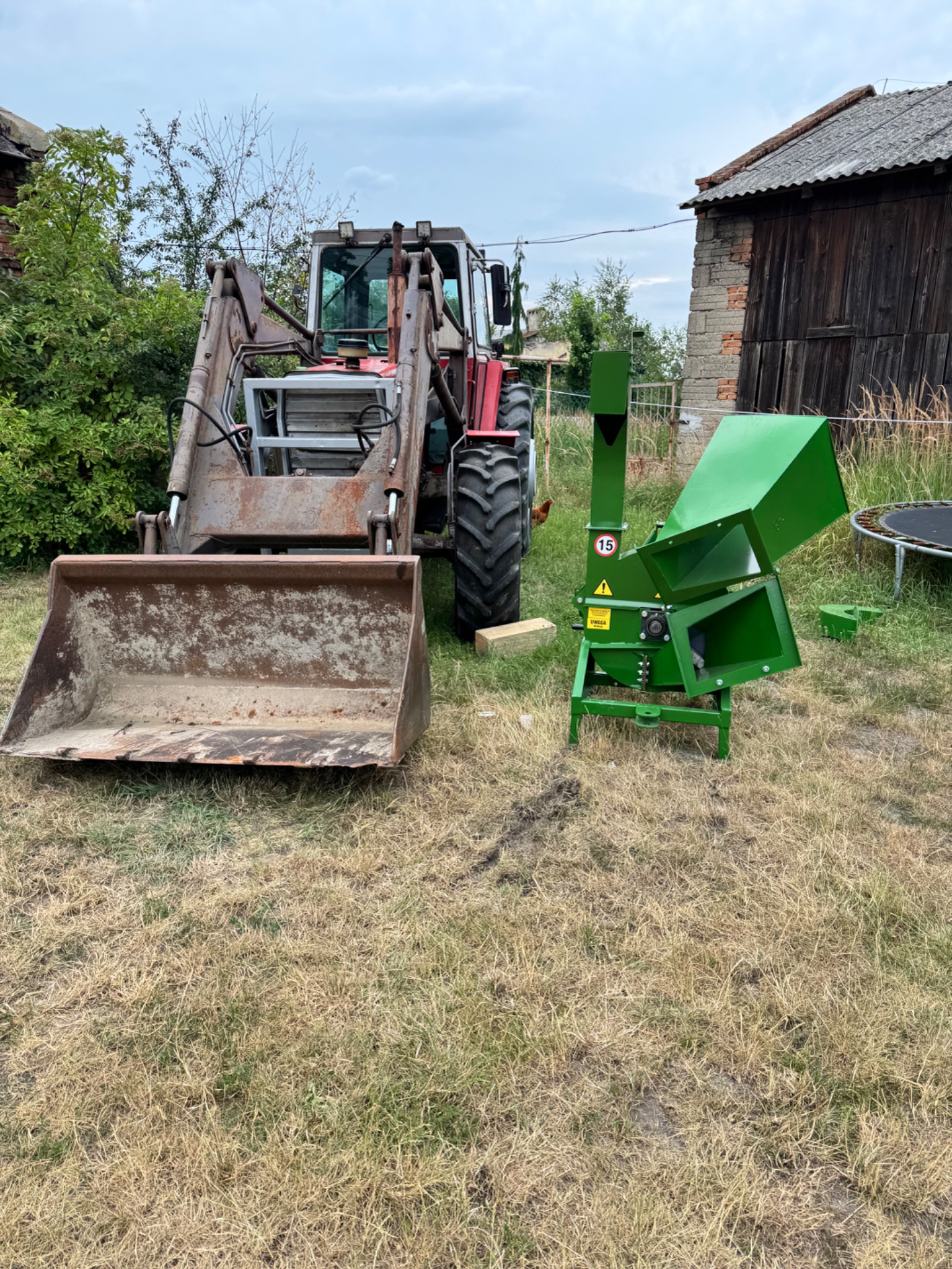 Traktor z łyżką i zielony rozdrabniacz do gałęzi na wyschniętej trawie, w tle budynek gospodarczy i drzewa.