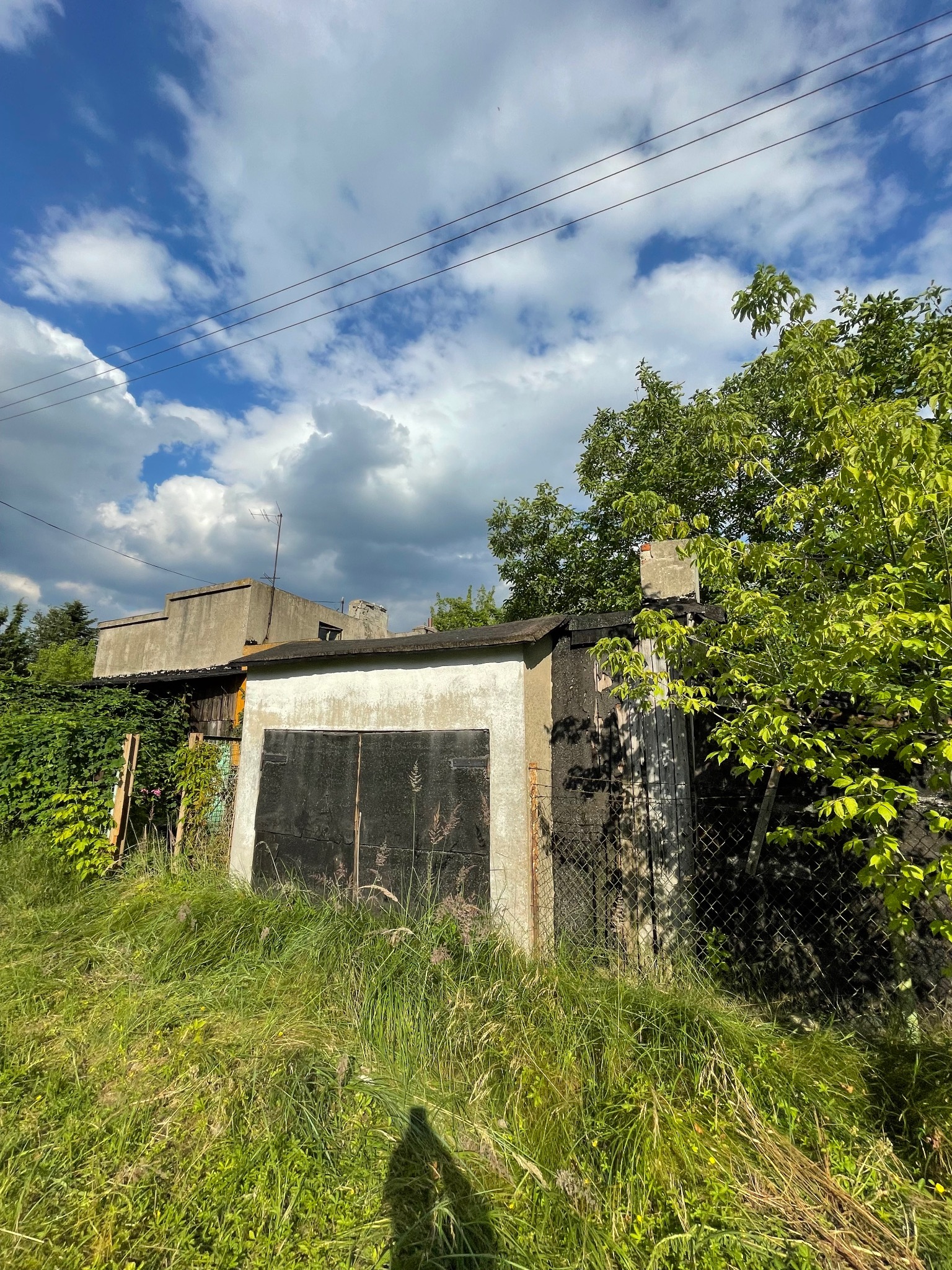 Stary, betonowy garaż z zardzewiałymi, metalowymi drzwiami, porośnięty trawą i krzewami, na tle błękitnego nieba z chmurami.