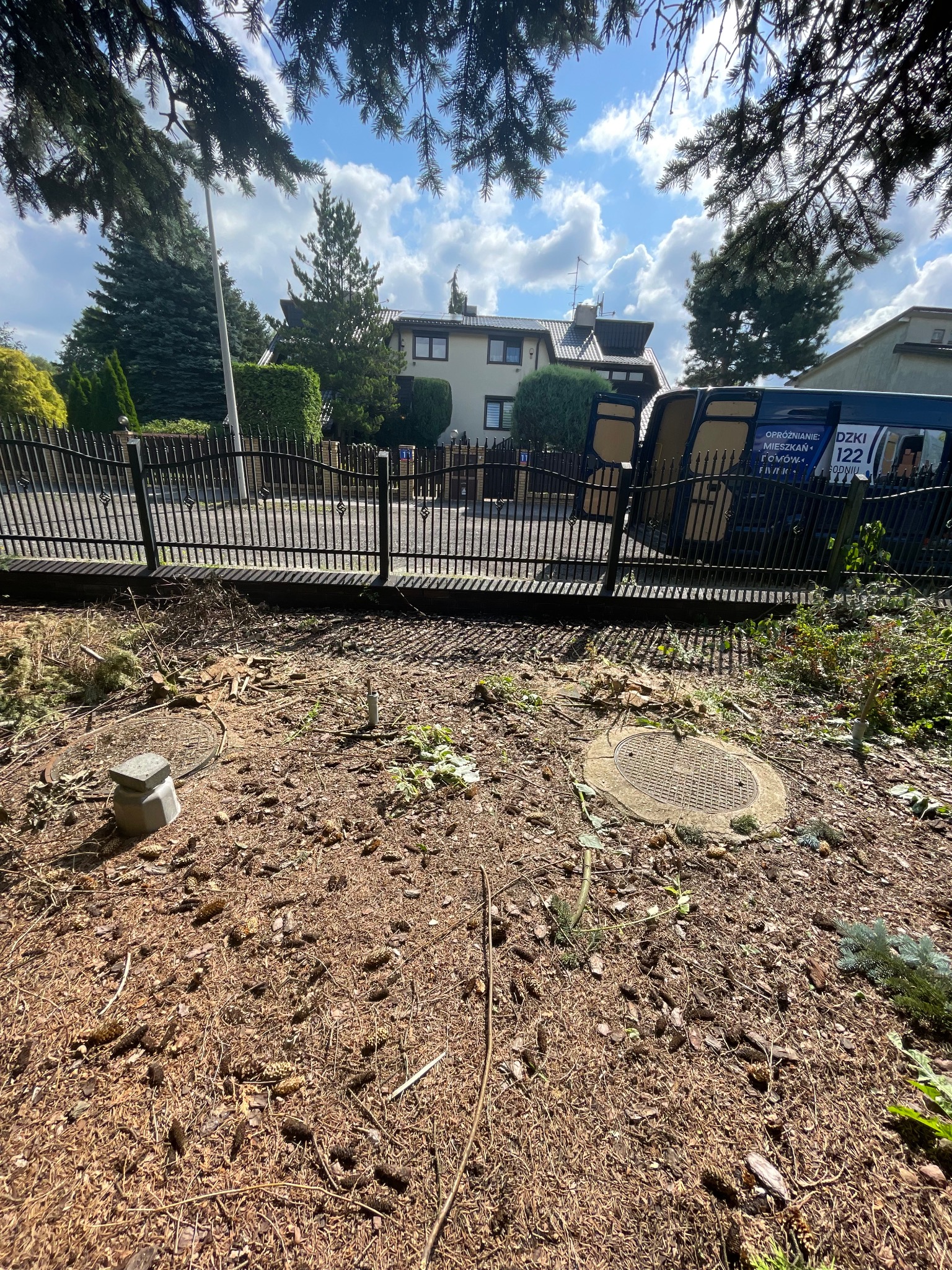 Teren po wycince drzewa z widocznymi pozostałościami gałęzi i pni, na tle ogrodzenia i zaparkowanego busa z reklamą.